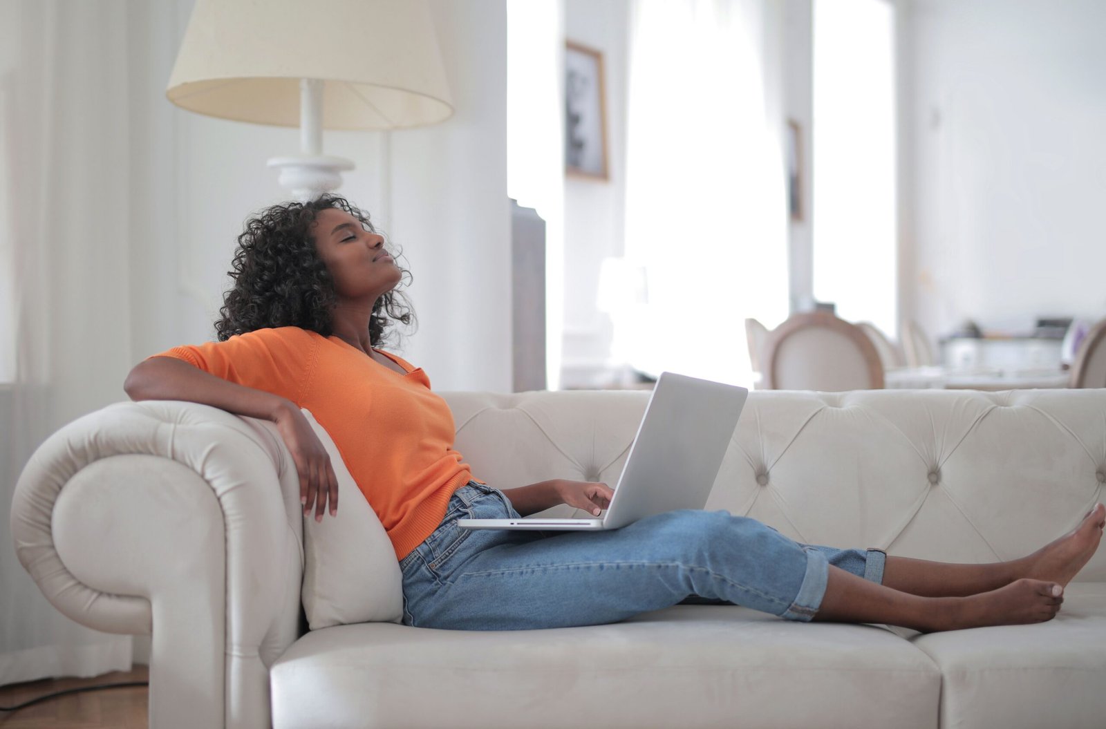 Woman in orange top relaxing on sofa with laptop, enjoying downtime.