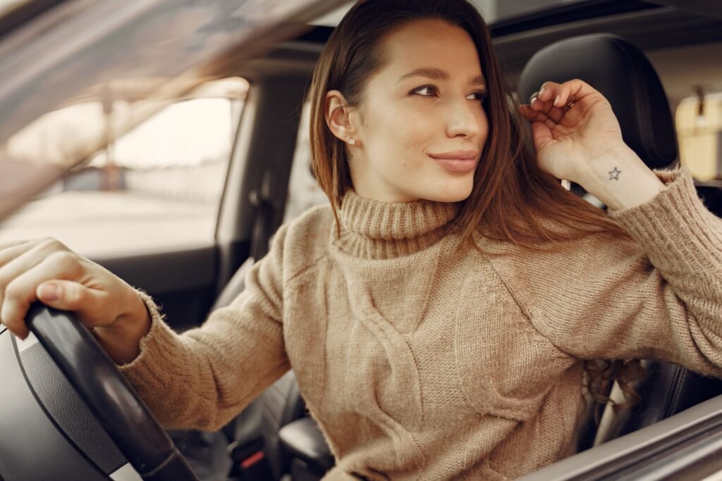 Positive young female driver in warm clothes at wheel of leather interior car going around city streets while greeting friend during riding